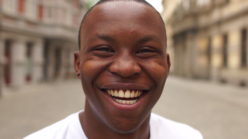 Portrait of black man very happy, smiling in urban background. An african american student sincerely smiles while standing in the middle of a city street. The guy is wearing a white t-shirt Royalty-Free Stock Footage #1054497092