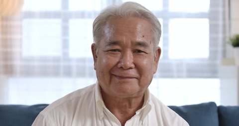 Closeup face portrait of healthy cheerful asian senior elderly old gray-haired man smiling to the camera while sitting on the blue couch in the living room. happy mature middle aged grandfather.