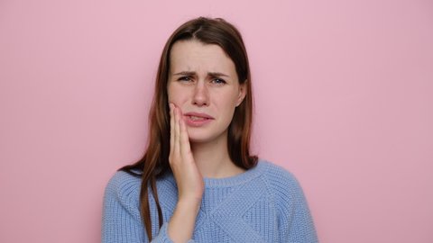 Unhappy young woman touch cheek, suffers from toothache, girl having terrible sudden tooth pain, need dental treatment, wears sweater, isolated on pink studio background. Medical insurance concept