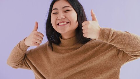 Asian teenager in brown pullover. She is smiling and showing thumbs up while posing against purple studio background. Close up