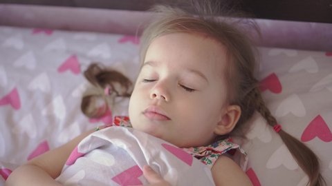 Sleep, rest, childhood, health concept - little blond-haired Slovenian Caucasian appearance child girl with two pigtails in a colorful dress wakes up smiling and stretches herself in bed