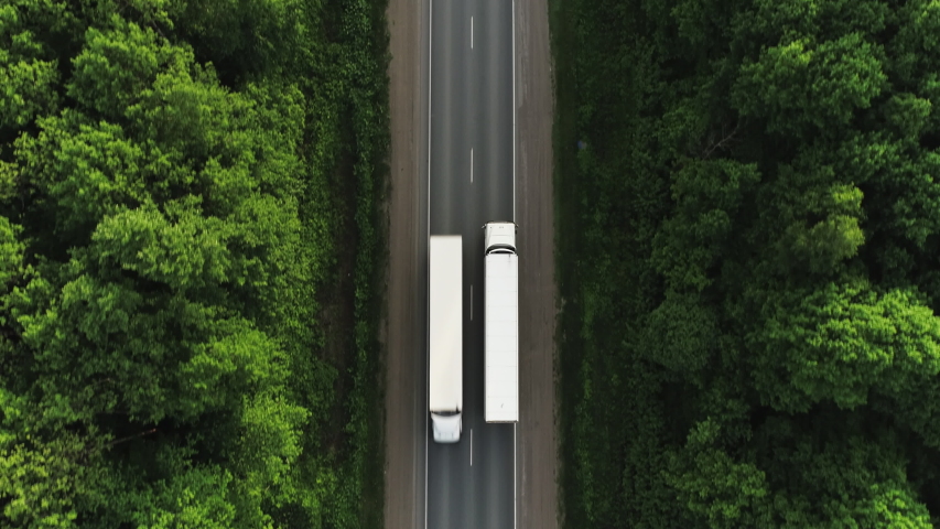 One Semi Truck with white trailer and cab driving / traveling alone on dense flat forest asphalt straight road, highway top down view follow vehicle aerial footage / Freeway trucks traffic