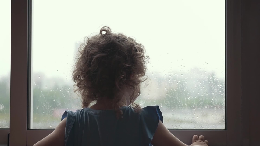 Silhouette of a little girl, which looking out the window on a rainy day. Child lonely standing beside window, and says goodbye to someone, waving her hand.  Royalty-Free Stock Footage #1054507835