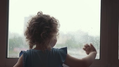 Silhouette of a little girl, which looking out the window on a rainy day. Child lonely standing beside window, and says goodbye to someone, waving her hand. 