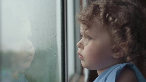 Sad little blond curly girl looks out the window on a rainy day. A child is waiting for her parents.