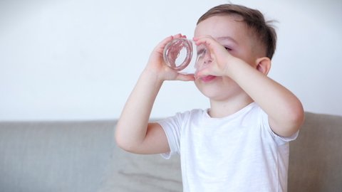 Cute baby boy drinking a glass of water sitting on the couch at home. Slow motion little boy drinking water. Close-up. Portrait Funny little Child is drinking a cup of water.