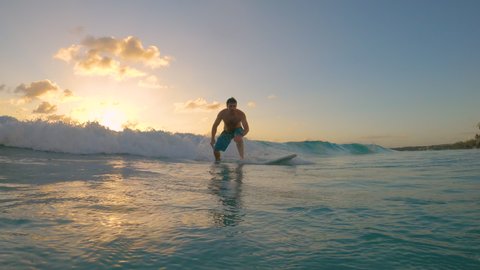 SLOW MOTION, LOW ANGLE: Cinematic shot of a happy male surfer riding a small wave to the tropical coast. Cheerful surfer dude on vacation in Barbados rides a small wave on a picturesque summer evening