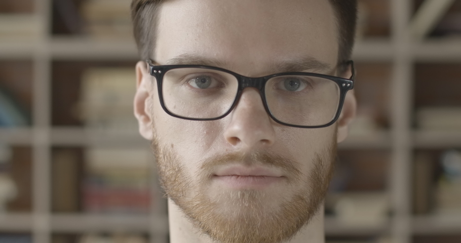 Extreme close-up face of handsome brunette man taking off eyeglasses in slow motion. Portrait of intelligent Caucasian guy with red beard looking at camera in library. Cinema 4k ProRes HQ.