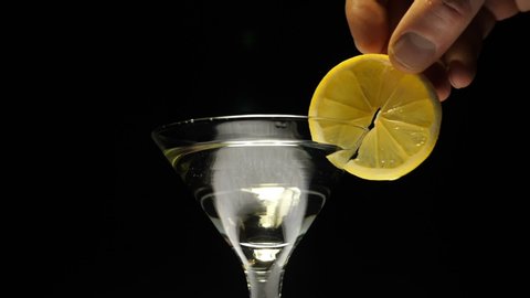A man pours a martini (white wine) from a bottle into a glass goblet and decoration with a slice of lemon on a black background. Closeup. The camera moves around. Slow motion. High speed camera
