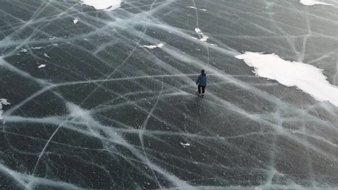 Aerial top view of a man ice skating outdoors on frozen water reservoir. Clip. Male tourist ice skating on frozen lake with thick ice with deeep cracks below his legs.