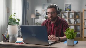 Young bearded man in red shirt at the desk in headphones speaks by video link in the living room