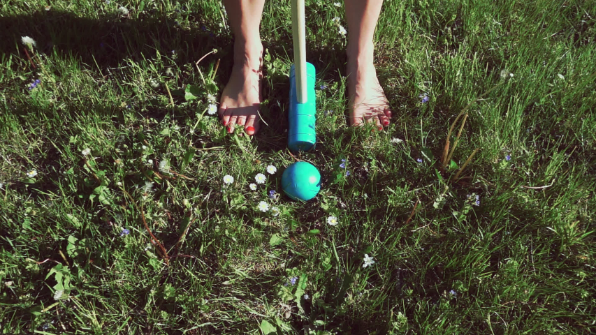 Woman striking a bright turquoise wooden ball in between her bare feet with a wooden mallet while playing garden croquet in slow motion.
