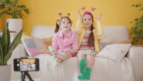 Shooting a Video Blog for Children with Smartphone. Production of Kid's Content. Two Cute Cheerful Little Caucasian Girl Talking and Laughing, Sitting on a Couch. Cozy Interior with Yellow Wall.