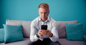Front view of bearded man with serious facial expression looking at smartphone screen while sitting on grey sofa. Young male in formal outfit using mobile during leisure time at home.