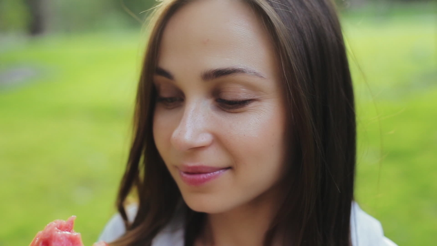 Close-up view of attractive cute brunette young woman with kind eyes, sniffing and eating slice of watermelon and smiling at the camera. Beautiful and pretty girl enjoy taste of fruit outside in park.