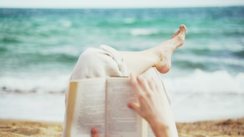 On the beach. A girl lies on the beach and reads a book. Female legs on the sea background. Light breeze. Turquoise waves. Summer chill. Vacation at the sea. Rest and pleasure. Holidays on the beach.