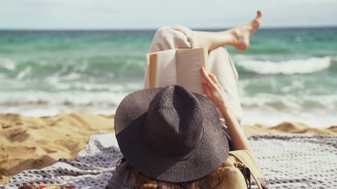 On the beach. A girl in a black hat lies on the beach and reads a book. Light breeze. Turquoise waves in the background. Summer chill. Vacation at the sea. Rest and pleasure. Holidays on the beach.