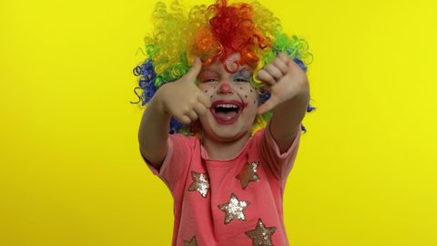 Little caucasian child girl in clown cosplay making silly faces. Colorful wig. Happy five years old female kid having fun, smiling. Thumb up, thumb down. Halloween. Isolated on yellow background