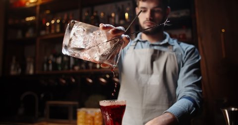 Authentic professional caucasian bartender creating a cocktail drink. Experienced barman pouring alcohol beverage in glass before serving - food and drink concept close up 4k footage