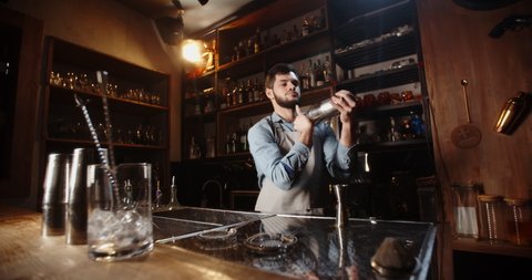 Cool professional caucasian bartender making a cocktail, shaking his shaker. Authentic barman making alcohol beverages in modern bar - food and drink 4k footage