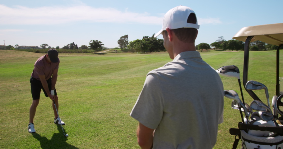 Caucasian male golfers standing on a golf course wearing golf clothes, one swinging a golf club and hitting the ball, while his opponent stands nearby in the foreground, with clubs in a golf buggy Royalty-Free Stock Footage #1054550816