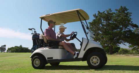 Two Caucasian male golfers on a golf course on a sunny day wearing caps and golf clothes playing a game, sitting in a golf buggy and driving off across the course, in slow motion 