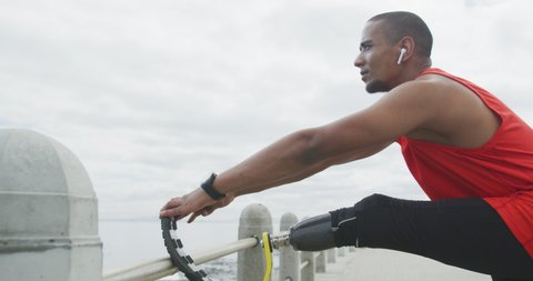 Side view close up of a disabled mixed race man with a prosthetic leg, working out on a promenade, stretching his disabled leg, leaning it on a barrier and bowing to it, listening to music on earphone