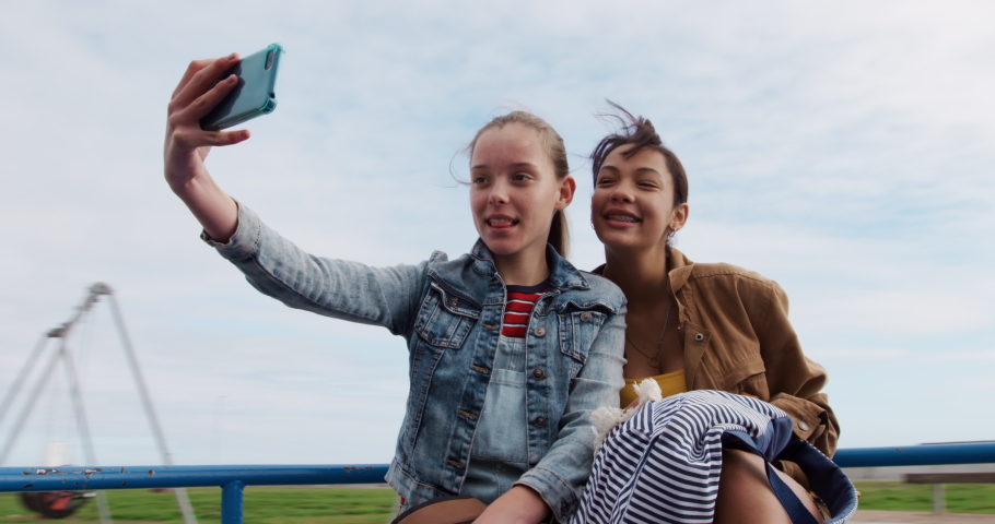 Front view of a Caucasian and a mixed race girl enjoying time hanging out together on a sunny day on a playground, sitting on a merry-go-round, smiling, girl taking selfie of herself and her friend