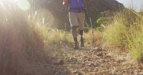 Low section of fit, disabled mixed race man with prosthetic leg, enjoying his time on a trip to the mountains, hiking, walking on the dirt road, using Nordic walking sticks, on a sunny day