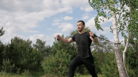 muscular man trains with elastic bands outdoor in park. Sportsman trains shoulders, arms and pectoral muscles.