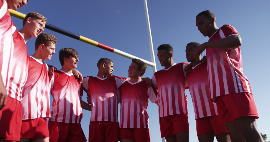 Low angle front view of a teenage multi-ethnic male team of rugby players wearing their team strip and standing on the playing field in a semi circle and arms around each other, talking in slow motion