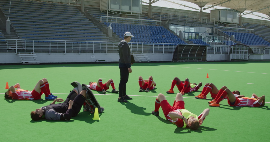 Side view of a multi-ethnic group of teenage male field hockey players and their Caucasian male field hokey coach training, doing sit ups in a circle while the coach stands watching, in slow motion Royalty-Free Stock Footage #1054559792