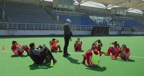 Side view of a multi-ethnic group of teenage male field hockey players and their Caucasian male field hokey coach training, doing sit ups in a circle while the coach stands watching, in slow motion
