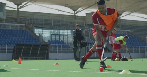 Side view of a mixed race male field hockey player, training before a game, running with a ball in a slalom holding a hockey stick, with the stadium and his teammates in the background, in slow motion
