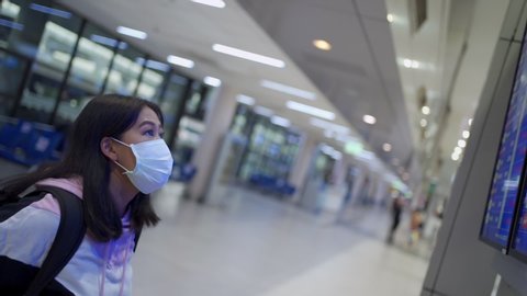 Asian girl wear protective mask standing and look at flight schedule time table screen, at airport terminal, on the move leaving hometown covid-19 pandemic, new normal, empty airport terminal