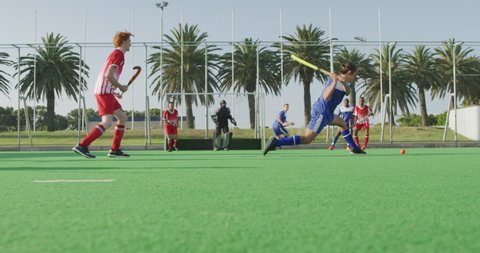 Side view of two multi-ethnic teenage male field hockey teams, playing a field hockey game, with one of the players hitting the ball with a hockey stick and goalkeeper defending the shot with his leg