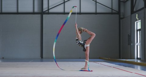 Side view of focused teenage Caucasian female gymnast performing at the gym, exercising with ribbon, spinning the ribbon, holding her leg behind her head, wearing multi coloured leotard