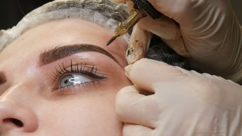 A special needle tattoo machine makes permanent makeup correction of a young woman's eyebrows. A pigment of dark paint is injected under the skin. Microblading, powder spraying.