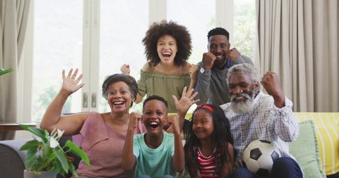 Multi-generation mixed race family at home, sitting on a couch, smiling, watching a game on tv, social distancing and self isolation in quarantine lockdown during coronavirus covid19 epidemic