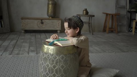 Cute muslim boy reading Koran in prayer hat and arabic clothes with rosary beads and Quran book praying to Allah, prophet Muhammad holy spirit religion symbol concept inside eastern interior
