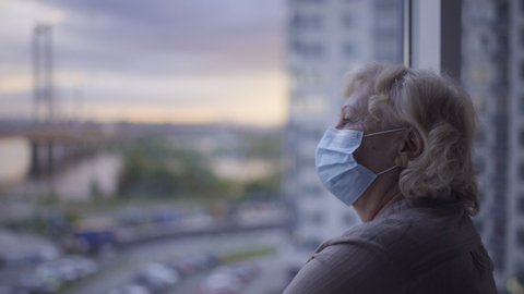Infected old patient standing by hospital window, wearing protective face mask