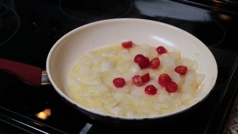 Male hand cooking white onions and grape tomatoes preparing food in a frying pan on a stovetop. Close-up of male hand stirring with a red spoon sauteing chopped onions and small grape tomato halves.