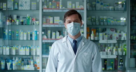 A pharmacist man stands in a pharmacy in a white uniform on the background of drugs and puts on a protective mask. Man portrait