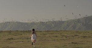 Young girl in white dress running to towards seagulls making them fly away, young woman running on green filed open enjoying. Freedom concept. birds fly in the sky. Wide frame slow motion. 