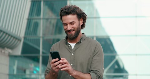 Attractive Handsome Man with nice Hairstyle and Beard Using his Modern Smartphone. Looking very stylish and Gorgeous. Wearing Khaki color Shirt and basic White Basic T-shirt. Typing Messages.