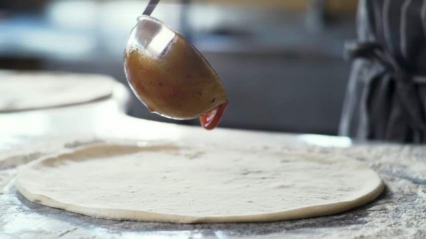 Close Up of Chef is spreading Tomato Sauce on Pizza Dough with a Metal Ladle in Traditional Italian Pizza's Restaurant. Cooking traditional Sauce for Italian Pizza. Italian Food. Pizza's Making. Royalty-Free Stock Footage #1054570112