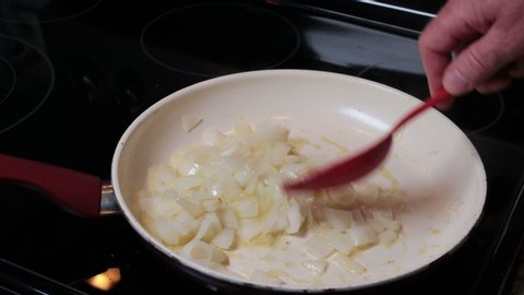 Male hand mixing while cooking sweet onions in a shallow ceramic frying pan on a black glass electric stove top. Close-up of a male hand stirring sauteing white onions in a pan on an electric stovetop