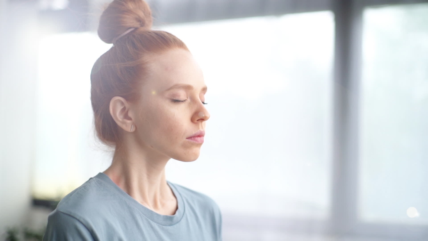 Close-up face of pretty redhead young woman doing breathing yogic practices at home office. Girl with closed eyes meditating on background of large window. Cute lady is making deep breath-exhalation. Royalty-Free Stock Footage #1054575686