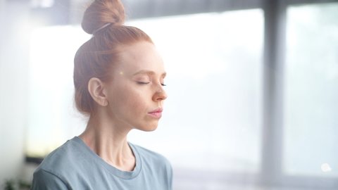 Close-up face of pretty redhead young woman doing breathing yogic practices at home office. Girl with closed eyes meditating on background of large window. Cute lady is making deep breath-exhalation.