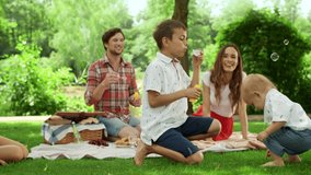 Happy man and woman spending weekend with children in park. Cheerful boys playing with soap bubbles outside. Joyful family having picnic outdoors. Smiling parents and kids sitting on blanket